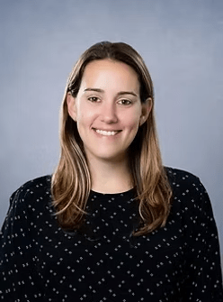 Woman with light brown hair smiles, wearing a dark top with white details, against a gray background.