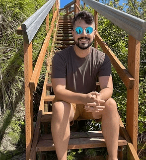 Man in sunglasses sits on wooden stairs outdoors, smiling. Brown shirt, khaki shorts.