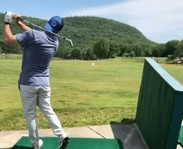 Golfer swings a club on a driving range, with green grass and a mountain backdrop.