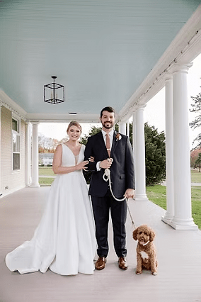 Bride and groom with a brown dog on a porch. The couple smiles, the groom holds the leash.