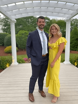 Man in suit and woman in yellow dress pose near a white pergola outdoors.
