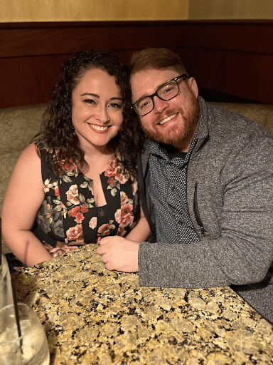 Couple smiling close together at a restaurant table. Woman wears floral dress, man in glasses and gray jacket.