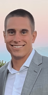 Man in a gray suit and white shirt smiles at the camera, against a soft-colored sky.