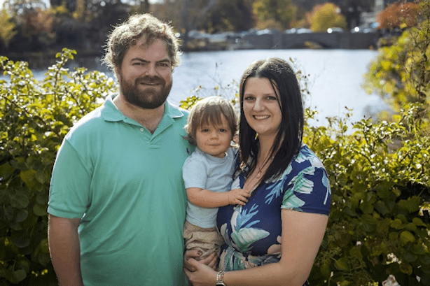 Family of three posing outdoors near water, man with beard, woman in floral print, holding toddler.
