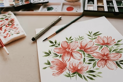 Watercolor painting of pink flowers and green leaves with art supplies.