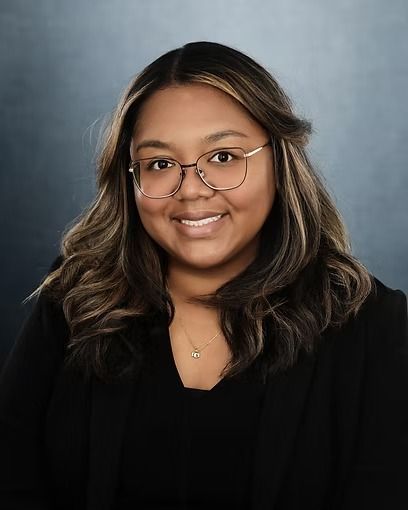 Woman with glasses smiles at the camera, wearing a black blazer. Against a blue-gray background.