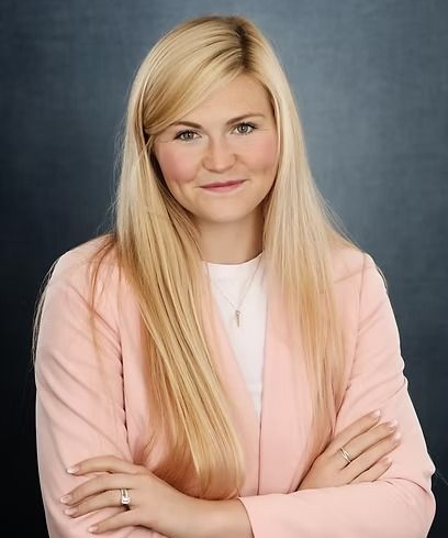 Blonde woman in pink blazer with arms crossed, smiling, against a dark blue background.