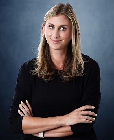 Woman with blonde hair, arms crossed, smiling, wearing black shirt. Blue background.