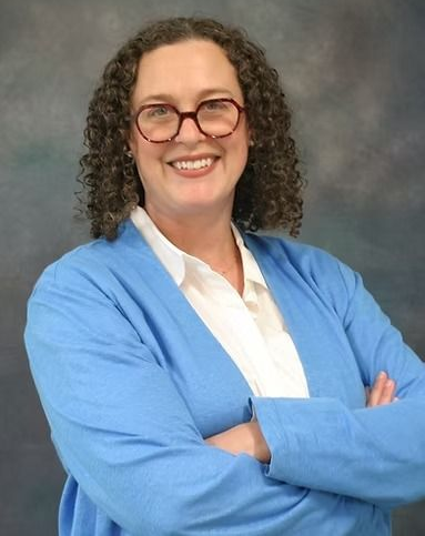 Woman with curly hair, glasses, and blue cardigan smiling, arms crossed.