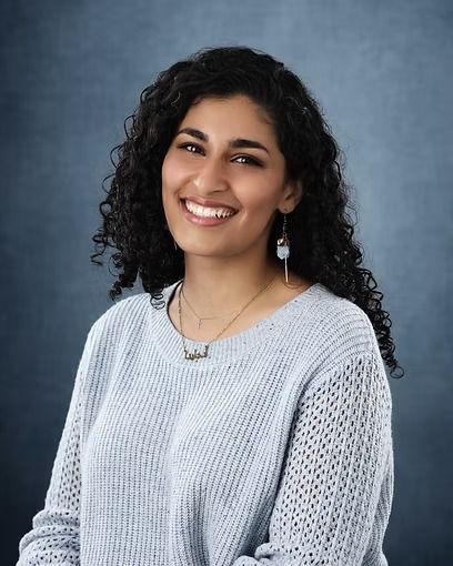 Woman with curly dark hair, smiling, wearing a blue sweater and necklace, against a blue backdrop.