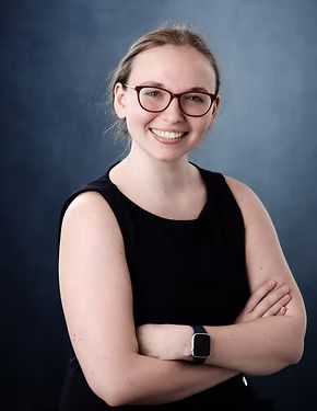 Woman with glasses and arms crossed, smiling. Dark sleeveless top, blue background.