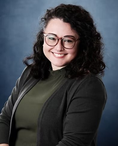 Woman with glasses smiles, wearing green shirt and gray cardigan, against a blue background.