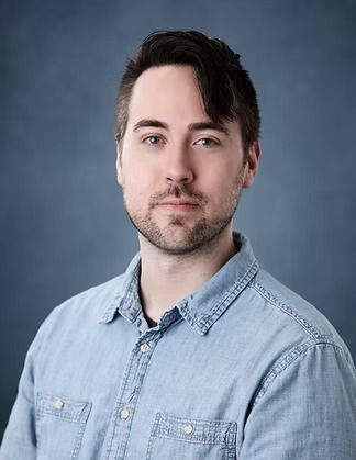 Man with brown hair and beard, wearing a denim shirt, looking at the camera against a blue backdrop.