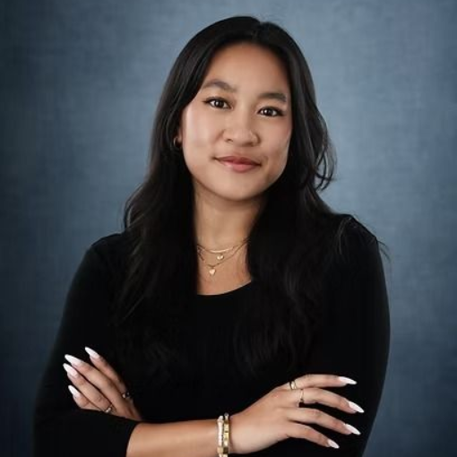 Woman with dark hair, crossed arms, wearing a black top and jewelry, smiling slightly against a blue backdrop.