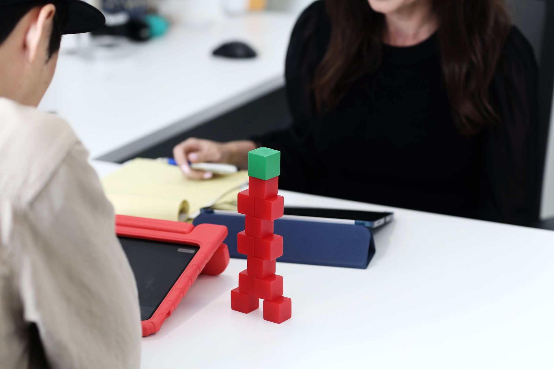 A person looks at a tower made of red and green blocks, with a second person seated behind them at a white table.