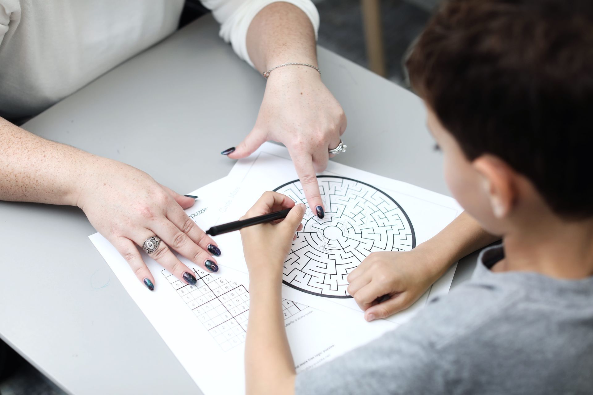 Person pointing at circular maze on a paper with another person. Both are working at a gray table indoors.