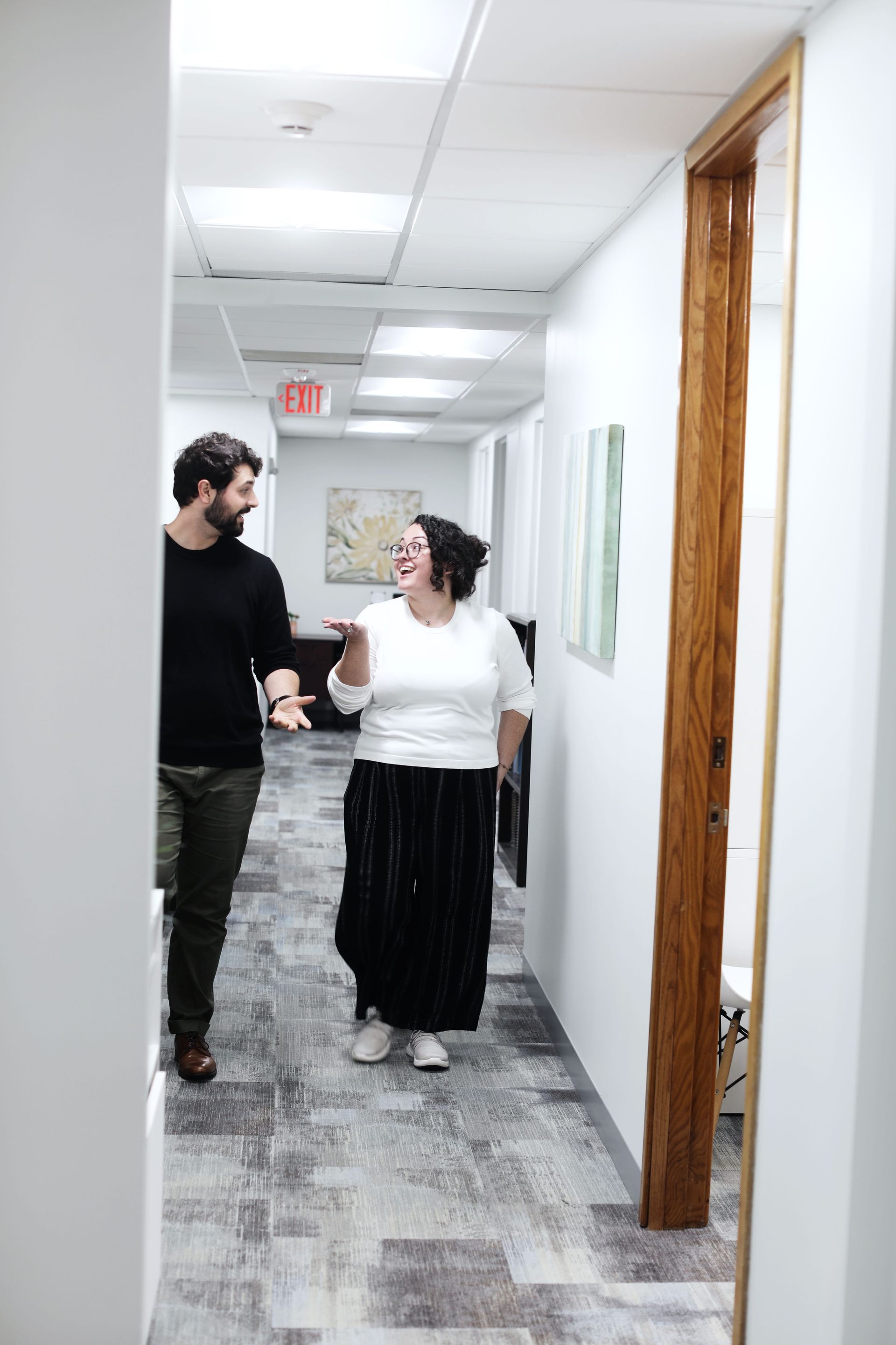 Two people smiling and walking down a hallway, white walls, carpet, artwork, open doorway.