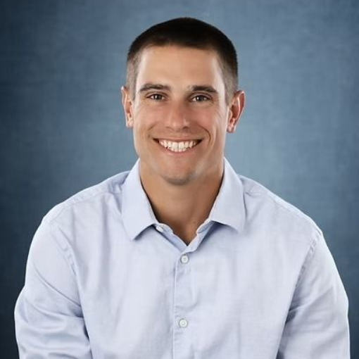 Man smiling in a light blue button-down shirt against a blue backdrop.
