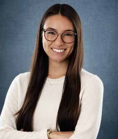Woman with long brown hair and glasses smiles. Wearing a white sweater, arms crossed, blue background.