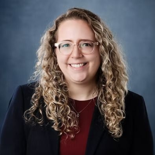 Woman with curly blonde hair and glasses smiles, wearing a blazer over a red shirt, against a blue background.
