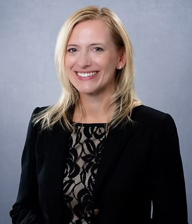 Woman with blonde hair wearing a black blazer smiles, looking at the camera.