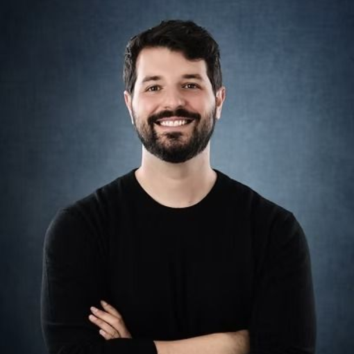 Man with a dark beard and black shirt smiles with arms crossed in front of a blue background.