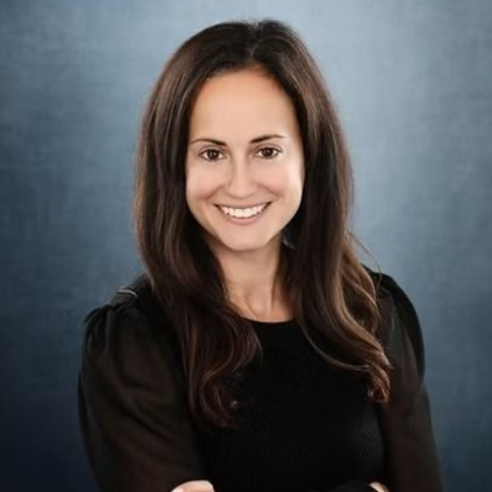 Woman with long brown hair smiling, wearing a black top against a blue-gray background.