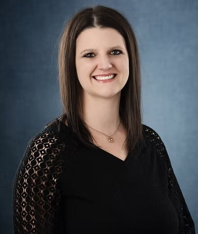 Woman with shoulder-length brown hair smiles at the camera. She wears a black top with lace sleeves, on a blue background.