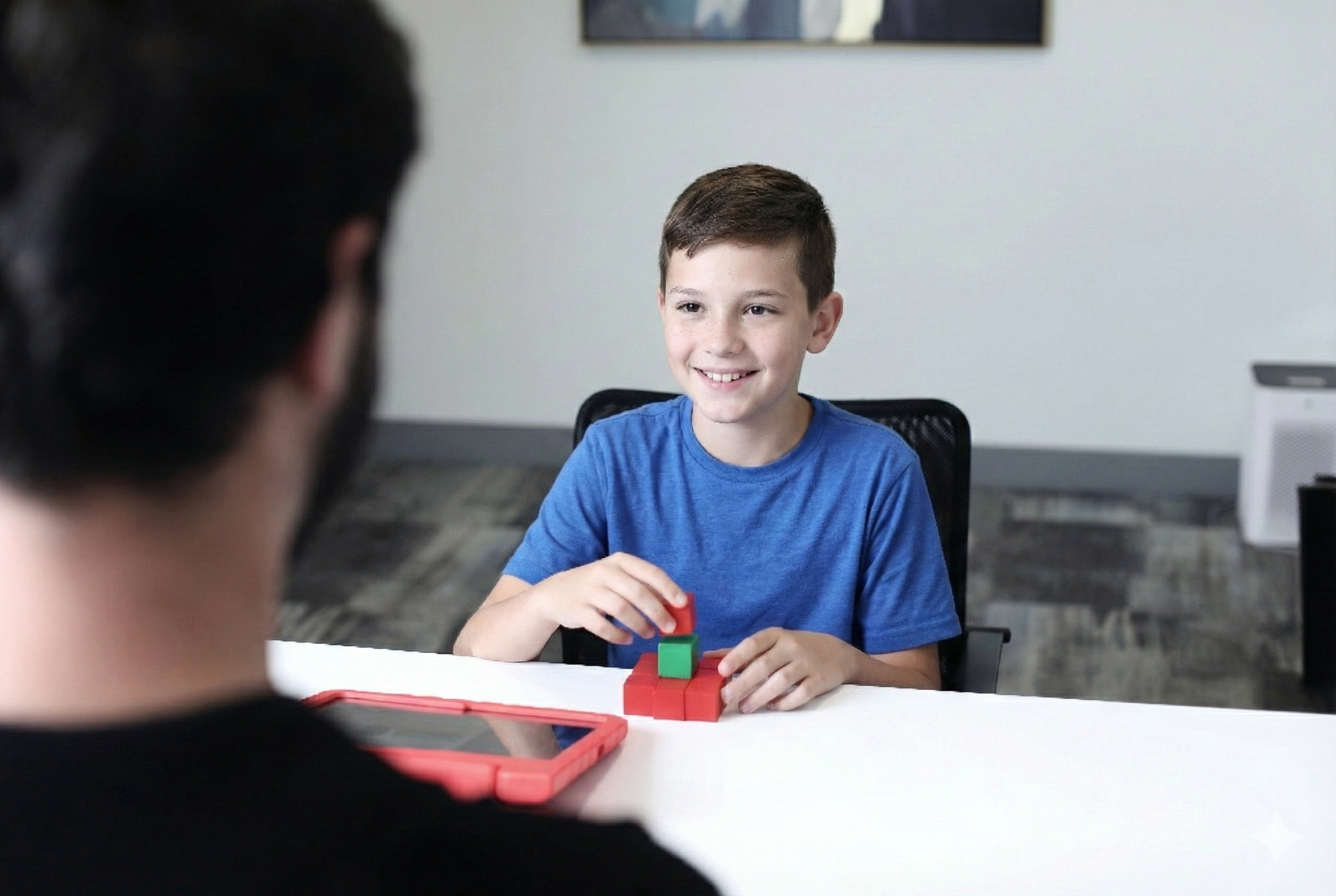 Boy playing with blocks at a table, smiling at person out of view. Red case tablet on the table.