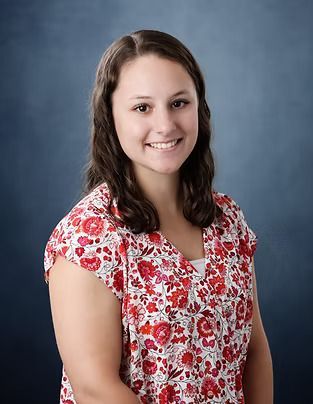 Woman with brown hair smiles, wearing a red and white floral top. Blue background.