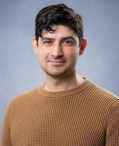 Man with dark hair and a brown sweater smiling, against a blue-gray background.