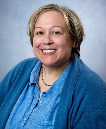 Woman with short blonde hair smiles, wearing a blue cardigan over a button-down shirt, with a neutral background.