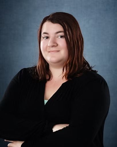 Woman with brown hair and arms crossed, wearing a black top against a blue backdrop.
