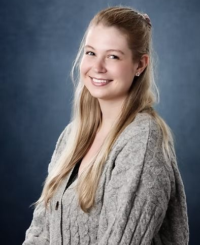 Woman with blonde hair, smiling, wearing a gray cable-knit sweater, standing in front of a blue backdrop.