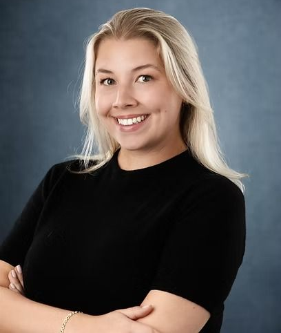 Blonde woman smiling, arms crossed, wearing a black shirt, set against a dark blue background.