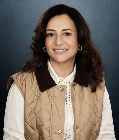 Woman in tan vest smiles at the camera, dark hair and hoop earrings, blue background.