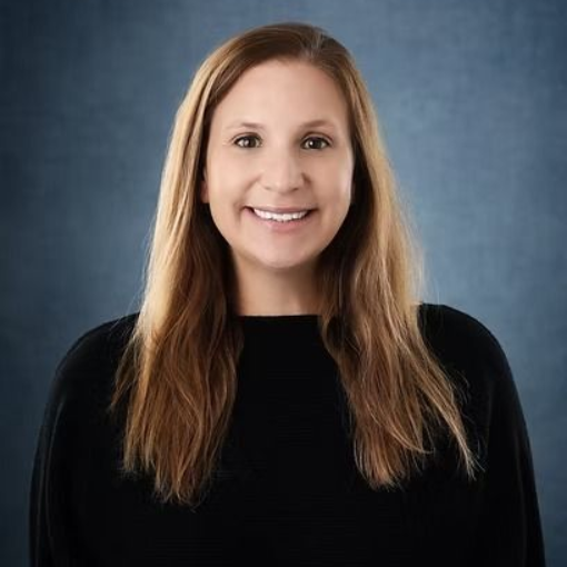 Woman with long brown hair smiles at the camera, wearing a black top. Blue backdrop.