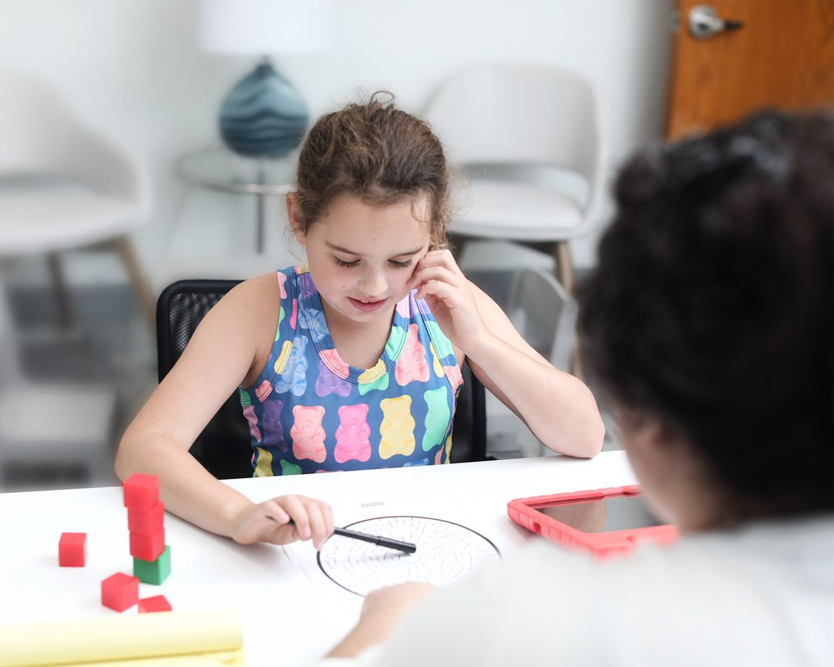 Girl drawing on paper at table, with person looking on. Red and green blocks are visible.