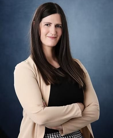Woman with long dark hair, arms crossed, wearing a tan blazer over a black top, against a blue background.