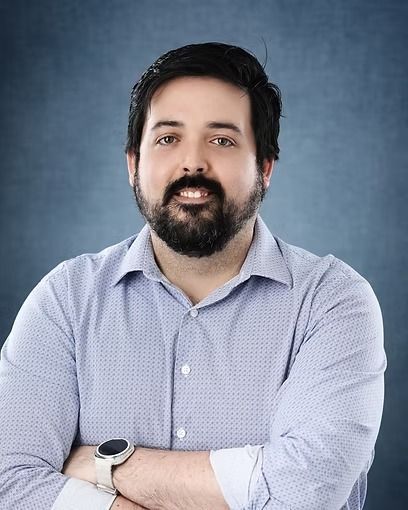 Man with dark hair and beard, arms crossed, wearing a light blue button-down shirt.