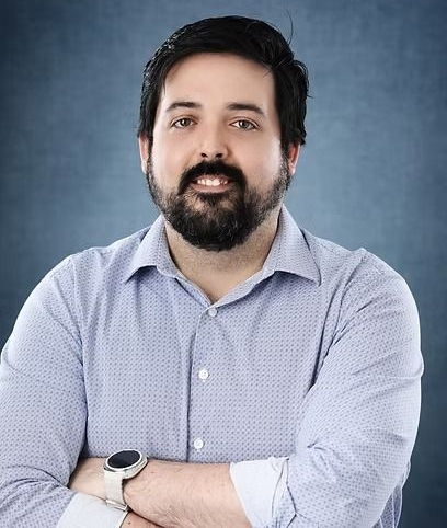 Man with dark hair and beard, arms crossed, wearing a light blue button-down shirt and a smartwatch.