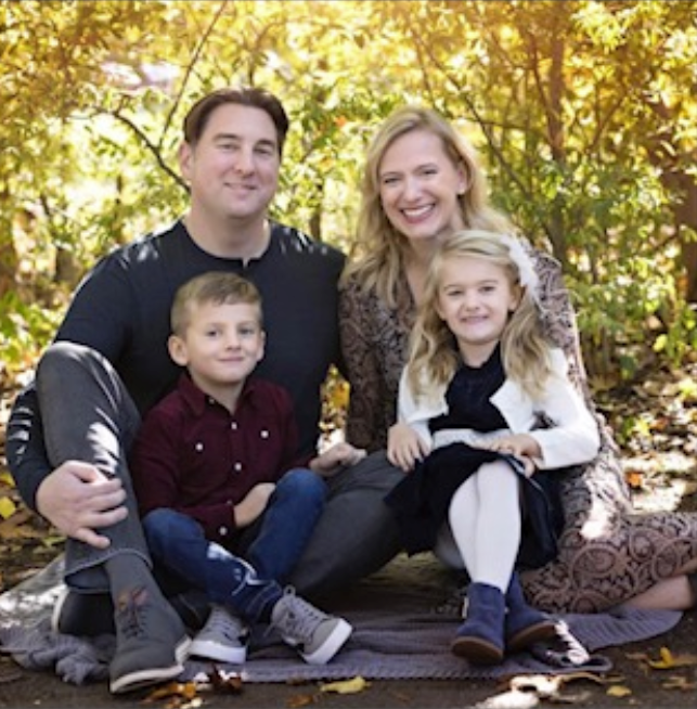 Man with dark hair and stubble in a denim shirt, looking directly at the camera against a blue backdrop.