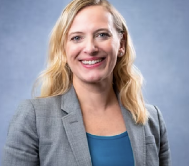 Woman with glasses smiles, arms crossed, wearing a black top against a blue background.
