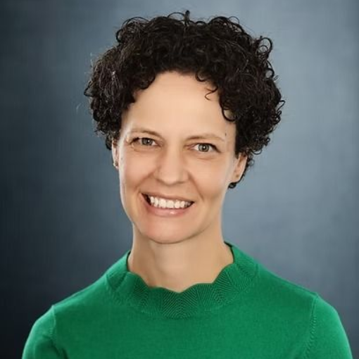Woman with curly dark hair, wearing a green sweater, smiling, against a blurred blue and gray backdrop.