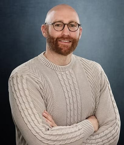 Bald man with a red beard wearing glasses and a light sweater, arms crossed, smiling.