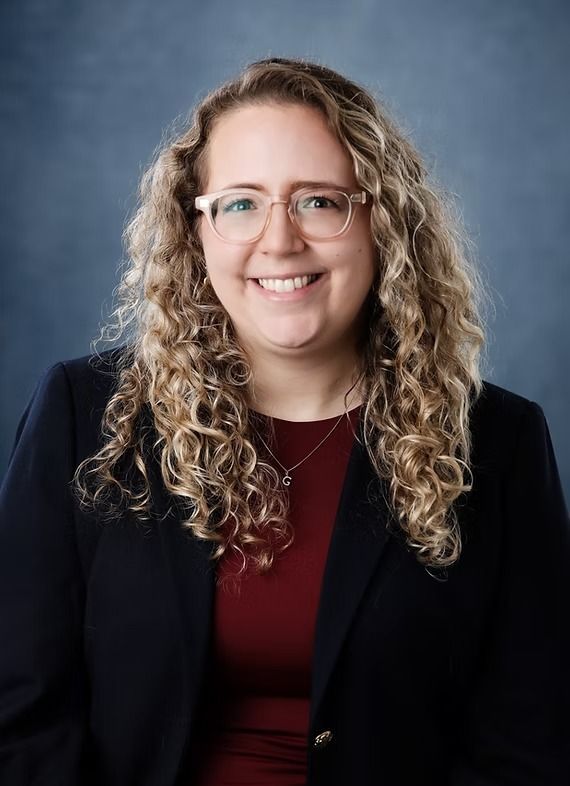 Woman with curly hair and glasses smiling, wearing a dark blue blazer and red shirt, on a blue background.