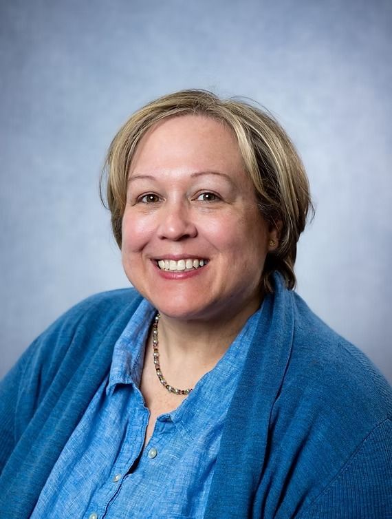 Woman with blonde hair smiling, wearing a blue shirt and cardigan, neutral background.