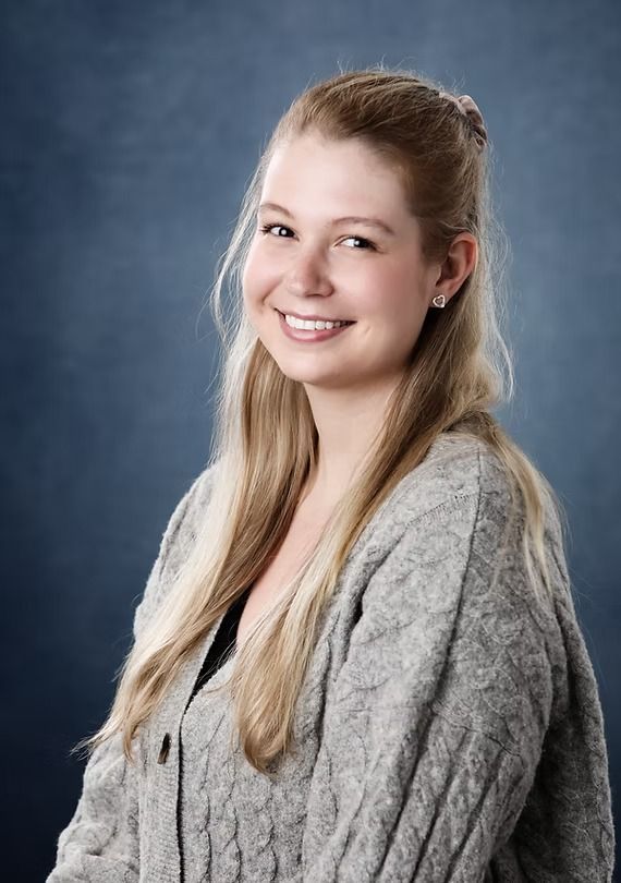Woman with blonde hair wearing a grey cable knit sweater, smiling against a blue background.