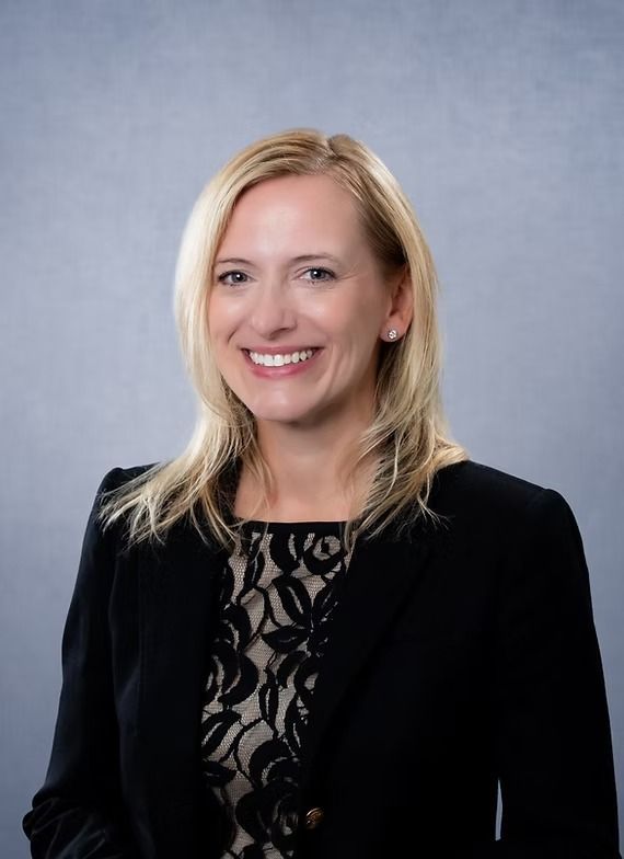 Woman with blonde hair wearing a black blazer smiles, posing in front of a gray backdrop.