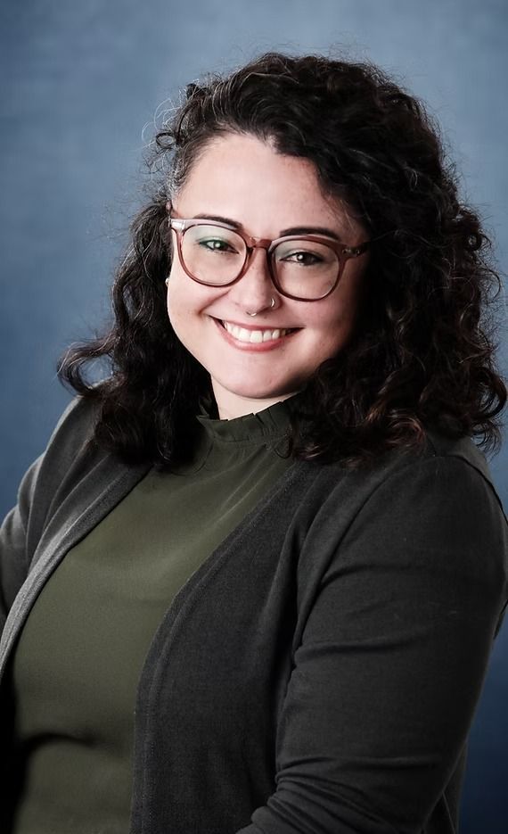 Woman with glasses smiles; dark curly hair, olive shirt, gray cardigan, blue background.